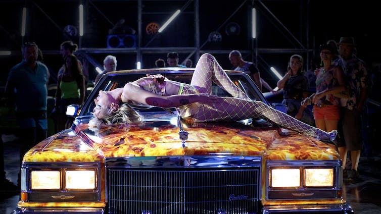 Una mujer en sujetador y medias de rejilla se tumba sobre el capó de un coche en un mundo futurista.